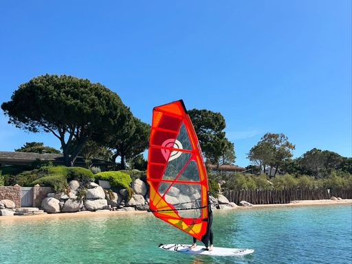 Planchiste à la voile rouge naviguant sur l'eau turquoise devant la côte de Cala Rossa